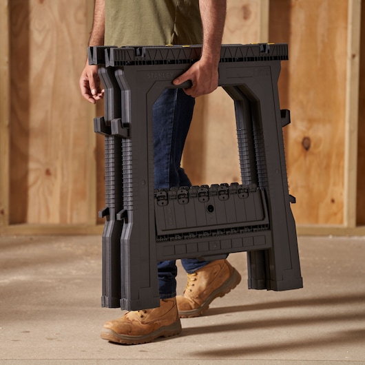STANLEY 060864R folding saw horse, dark gray, shown being carried by a person, product photography.