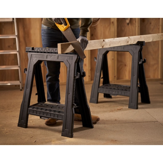 STANLEY 060864R black plastic sawhorse pair holding wood in workshop, side angle.