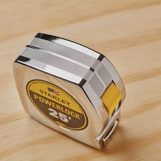 STANLEY 33-425 tape measure with chrome case, yellow lock button, and 25 ft length, angled on wood surface.