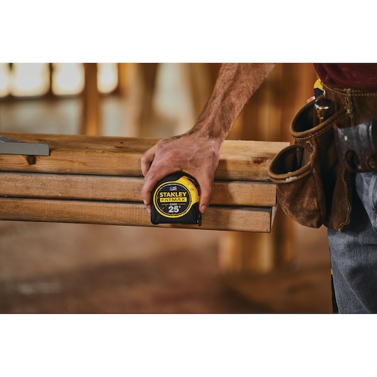 Photo of STANLEY 33-725 tape measure held by a person measuring wooden beams in a construction setting.