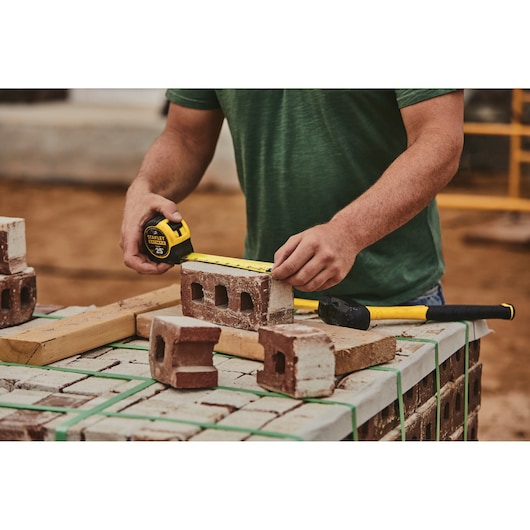 STANLEY 33-725 tape measure in use measuring a brick at a construction site, bricks and hammer visible on table.