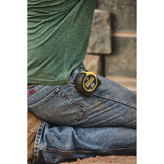 STANLEY 33-725 tape measure clipped to jeans pocket, person kneeling outdoors, product in use.