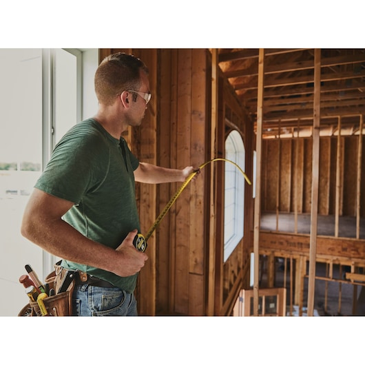 STANLEY 33-725 tape measure in use at a construction site, held by a person measuring a wooden frame wall.