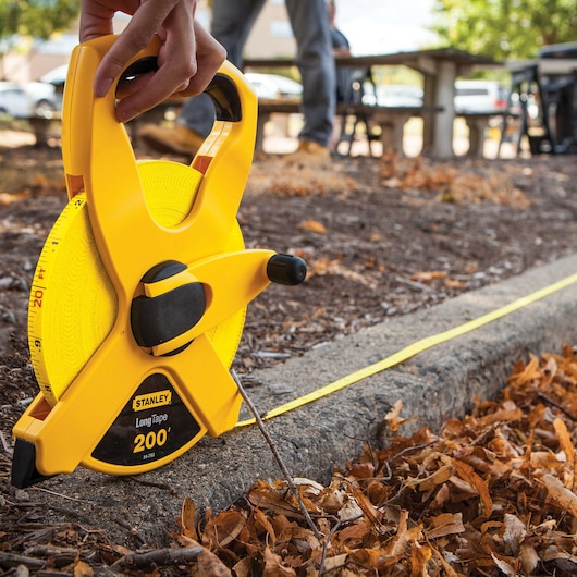 STANLEY 34-793 long tape measure, yellow, in outdoor use along a curb with fallen leaves visible.