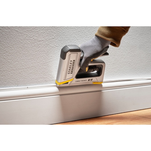 Photo of STANLEY FMHT70100 cable tacker securing wire along baseboard, side angle, gloved hand operating tool.