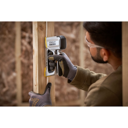 Stanley FMHT70100 FATMAX stud finder in use against wooden framing, user wearing gloves.