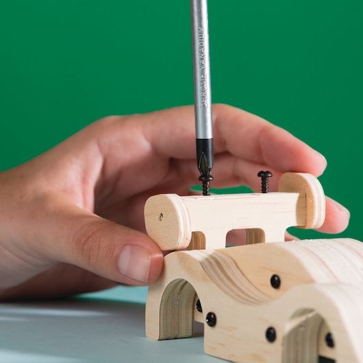 STANLEY JK030-SY screwdriver drives screw into wooden toy, close-up detail against green background.