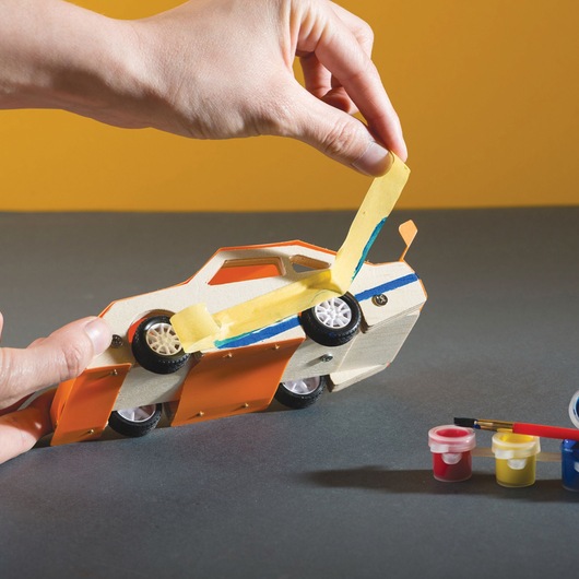 STANLEY OK002-SY photography of a hand peeling tape from a painted wooden toy car, paint pots and brush nearby.