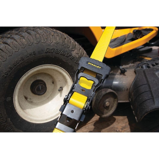 Photo of STANLEY S1007 ratchet strap securing yellow equipment near a lawn mower tire.
