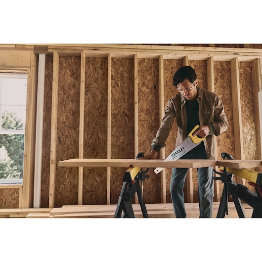 STANLEY STHT20350 hand saw cutting wood on sawhorses inside a framed construction area.