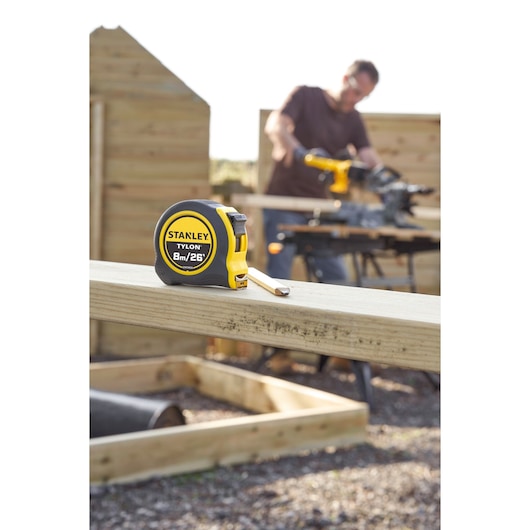 Photo of STANLEY STHT39526 tape measure on wooden beam outdoors, yellow and black case, 8m/26ft marking visible.
