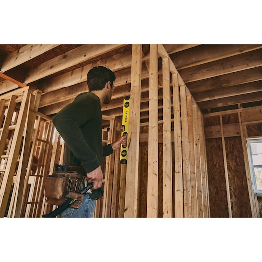 STANLEY STHT42496 level held by a person checking vertical alignment of wall studs in a wooden frame construction.