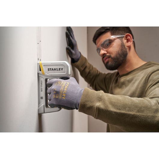 Photo of STANLEY STHT70150 staple gun in use, held by a gloved hand against a wall, side angle.