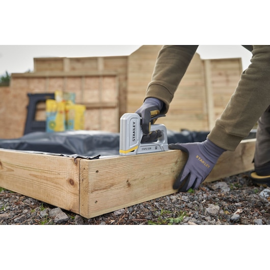STANLEY STHT70150 staple gun in use fastening plastic to a wooden garden bed, angled side view.