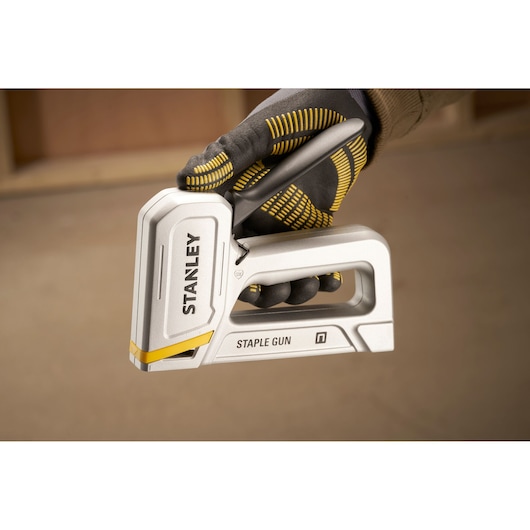 Stanley STHT70150 staple gun held by gloved hand, silver body, yellow accents, close-up view.