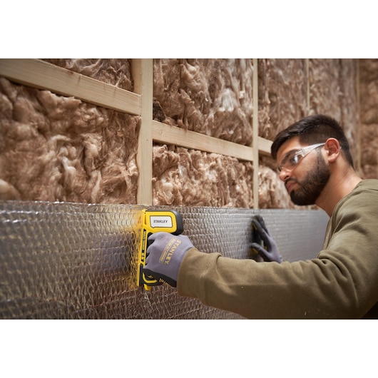 Stanley STHT70440 hand stapler used to fasten insulation material to a wall frame, person wearing work gloves.
