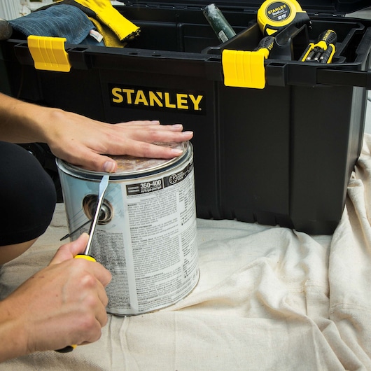 Photo of STANLEY STST33031 black tool box with yellow latches, paint can and hand using screwdriver.