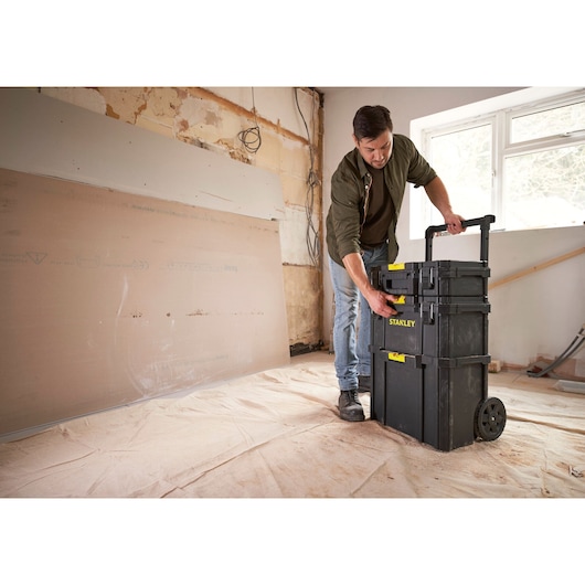 Photo of STANLEY STST60500 rolling tool storage box with yellow latches in a construction room, person adjusting handle.