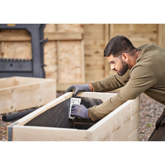 STANLEY TR45 stapler used to secure fabric inside a wooden garden bed, user wearing gloves outdoors.