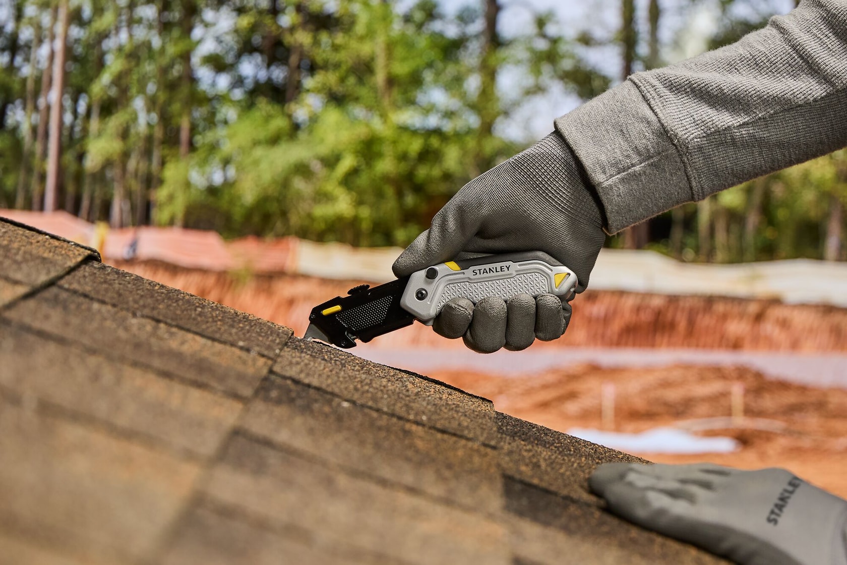 A person wearing work gloves is using a STANLEY utility knife to cut roofing shingles outdoors.