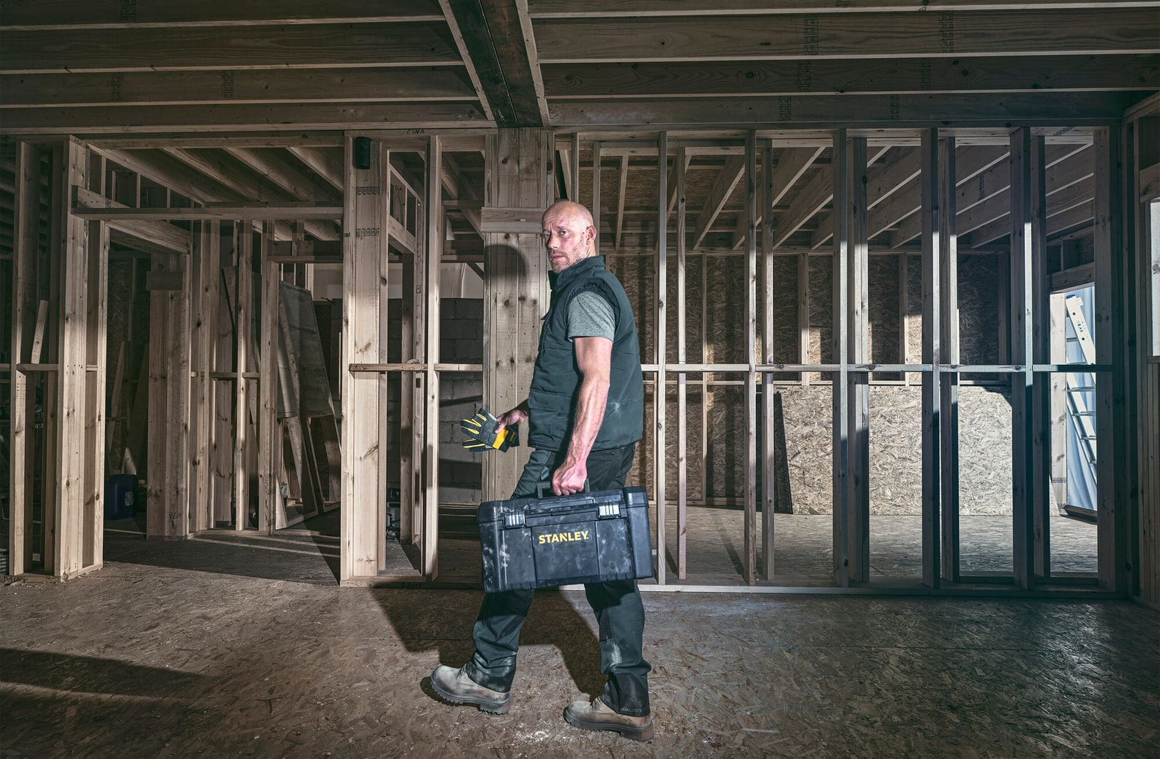 Photo of person holding STANLEY toolbox and tools inside wooden building framework, construction site setting.